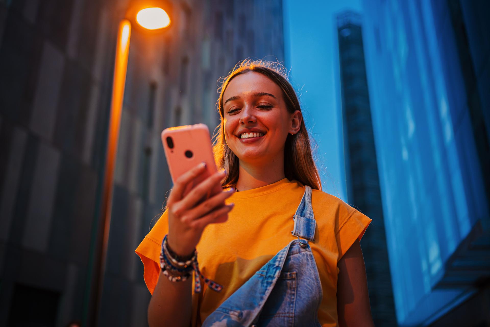 Real young adult woman using a cellphone at night. Happy female having fun with a smartphone social media app at dusk. Teenage girl browsing on internet with a mobile phone application at evening.